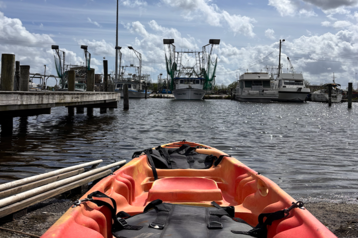 Orange kayak on shore facing a marina with boats under a cloudy sky.