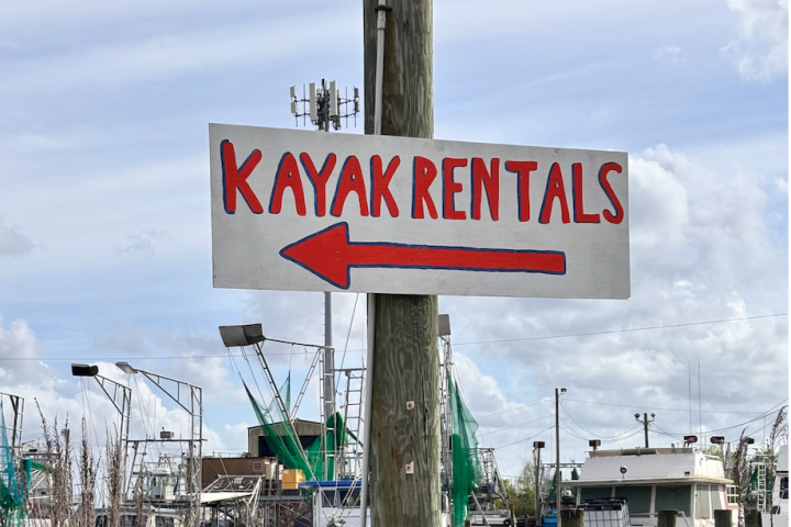 Sign reading 'Kayak Rentals' with arrow, near boats and cloudy sky.